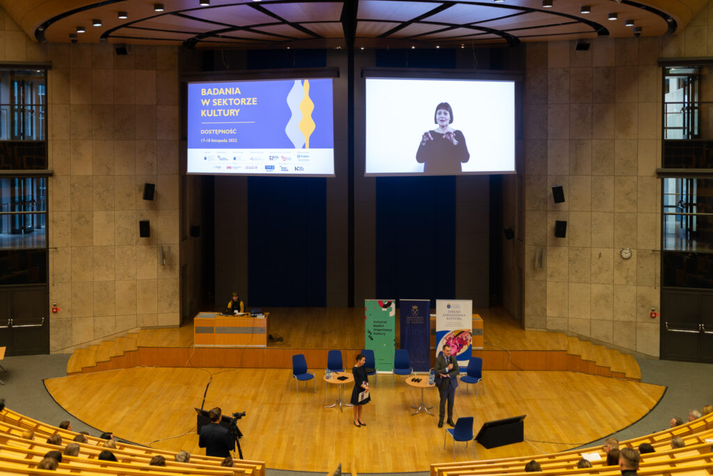 Rozpoczęcie konferencji - na scenie dwie osoby, nad nimi ekran z logotypem konferencji i tłumaczem języka migowego
